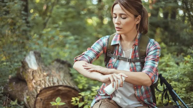junge frau sitzt im wald auf einem baumstamm und besprüht ihre nackten unterarme mit einem insektenschutzspray